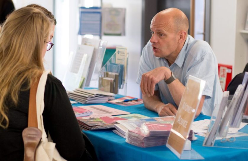 A student sits at a table talking to the university Money Adviser. A variety of leaflets are located on the table. 